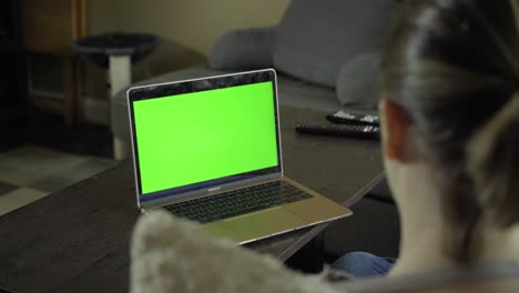 Over-the-shoulder-of-young-woman-sitting-on-a-sectional-couch-while-watching-green-screen-on-laptop-during-the-day-in-a-modern-home,-camera-pan-right