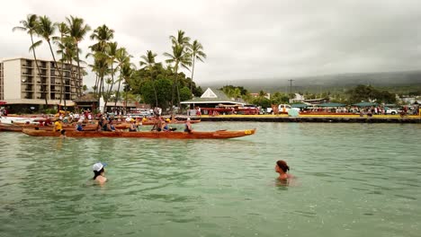 Two-young-ladies-swimming-conversing-in-bay-as-canoes-land-after-regatta-race,-Kona