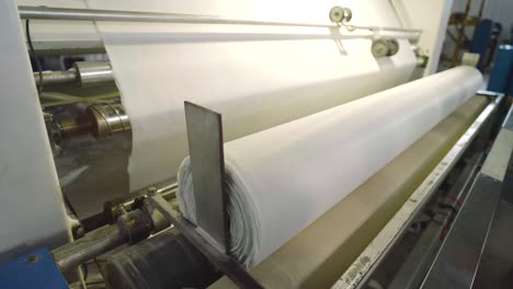 Large-White-Fabric-Being-Rolled-By-Machine-Tended-To-By-Worker-In-Textile-Factory