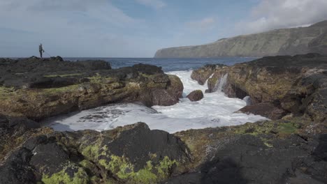 Man-stands-on-coastal-rocks-at-Mosteiros,-Sao-Miguel,-waves-crashing-through-natural-arches,-with-cliffs-in-the-background