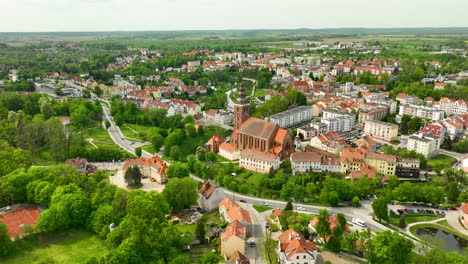 An-aerial-view-focusing-on-the-large-church-in-Lidzbark-Warmiński,-surrounded-by-residential-buildings-with-red-tiled-roofs-and-green-trees,-with-the-town-extending-into-the-background