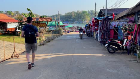 Young-blond-American-tourist-walking-through-a-local-clothing-market-in-the-rural-town-of-Sangkhlaburi-in-Kanchanaburi-province-on-a-sunny-afternoon,-Thailand