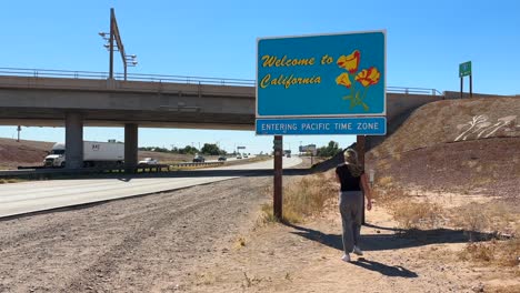 Mujer-Feliz-De-Pie-Con-El-Cartel-De-Bienvenida-A-California-En-El-Desierto