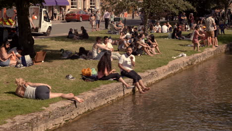 During-the-COVID-19-pandemic,-it-is-seen-people-are-enjoying-a-sunny-day-by-the-side-of-a-canal-in-Bourton-on-The-Water,-Cotswold,-United-Kingdom