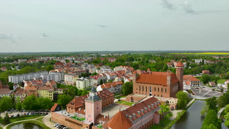 A-broader-aerial-view-of-Lidzbark-Warmiński,-showcasing-the-historic-castle,-surrounding-buildings-with-red-tiled-roofs,-and-green-trees,-with-more-of-the-town-in-the-background