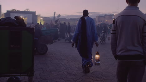 Tall,-dark-skinned-man-holding-flaming-oil-lantern-walks-through-Jemaa-El-Fna-medina-in-Marrakech,-Morocco