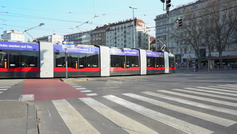 Una-Toma-Cámara-En-Mano-De-Una-Intersección-En-Viena-Con-Dos-Tranvías-Pasando-Y-Gente-Caminando-Sobre-Un-Paso-De-Peatones-Al-Fondo.