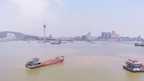 Hohe-Drohnenaufnahme-Eines-Lastwagens,-Der-An-Einem-Hopperkahn-Und-Einem-Kobelco-Kobe-Stahlgreiferbagger-Vorbeifährt,-Mit-Der-Skyline-Von-Macau-Im-Hintergrund