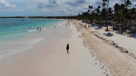 Woman-walking-on-beautiful-tropical-beach-with-palm-trees-in-the-Dominican-Republic,-aerial-descending-tilting-up-shot