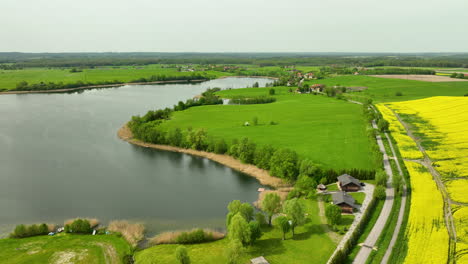 Luftaufnahme-Eines-Sees,-Umgeben-Von-Grünen-Feldern,-Kleinen-Häusern-Und-Einem-Rapsfeld,-Unter-Einem-Klaren-Himmel