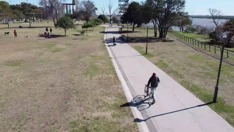 Hombre-Caminando-Junto-A-Su-Bicicleta-En-La-Orilla-Del-Río-Junto-A-Un-Parque-Donde-La-Gente-Se-Divierte