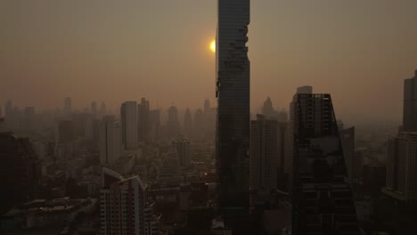 Aus-Der-Luftperspektive-Entfaltet-Sich-Die-Skyline-Von-Bangkok-Und-Präsentiert-Städtische-Wohnhäuser-Inmitten-Eines-Himmels,-In-Dem-Die-Aufgehende-Sonne-Durch-Neblige-Und-Rauchige-Schichten-Bricht-Und-Eine-Stimmungsvolle-Szene-Schafft