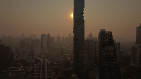 Desde-Una-Perspectiva-Aérea,-El-Horizonte-De-Bangkok-Se-Despliega,-Mostrando-Residencias-Urbanas-En-Medio-De-Un-Cielo-Donde-El-Sol-Naciente-Atraviesa-Capas-De-Niebla-Y-Humo,-Creando-Una-Escena-Atmosférica.