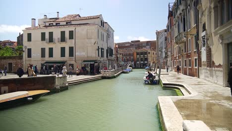 Vista-Panorámica-De-Un-Canal-Con-Edificios-Arquitectónicos-Italianos-Y-Turistas-Caminando-Por-Venecia.