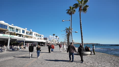 People-stroll-along-a-sunny-waterfront-promenade-lined-with-palm-trees-and-cafes-in-Pafos,-Cyprus