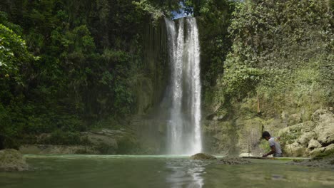 En-Las-Cataratas-Camugao,-En-Filipinas,-El-Agua-Cae-En-Cascada-Mientras-Un-Artesano-Local-Trabaja-Diligentemente-En-Su-Tabla-De-Surf,-Una-Vívida-Ilustración-De-Viajes-Y-Exploración.