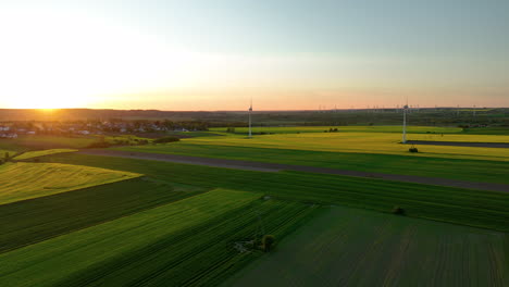 Luftaufnahme-Von-Grünen-Feldern-Und-Einem-Entfernten-Dorf-Bei-Sonnenuntergang-Mit-Windturbinen