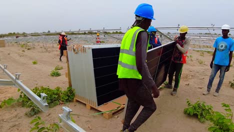 Imagen-Estática-En-Cámara-Lenta-De-Trabajadores-Africanos-De-La-Construcción-Usando-Cascos-Mientras-Levantan-Paneles-Fotovoltaicos-Para-Obtener-Energía-Sostenible-Y-Protección-Ambiental-En-Gambia,-África-Occidental