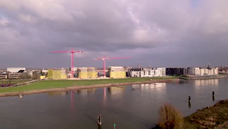 Aerial-River-IJssel-passing-by-Dutch-new-build-neighbourhood-Noorderhaven-with-red-cranes-rising-above-construction-site