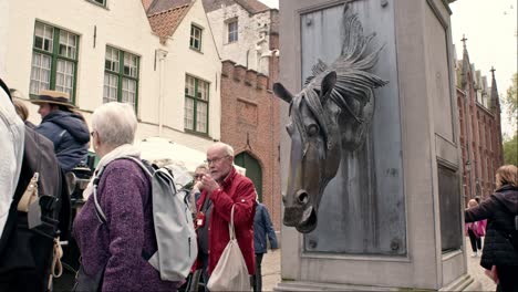 Closeup-Of-Horse-Head-Drinking-Fountain-With-Old-People-Walking-In-Bruges,-Belgium