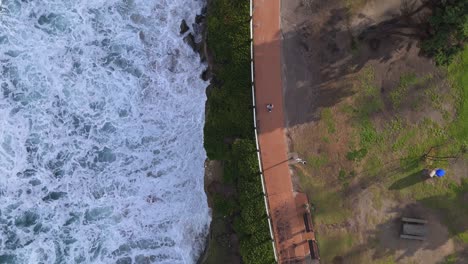 La-Jolla-Cove-Drone-Top-Down-Shot-Split-over-waves-crashing-while-person-walks-along-park-path-from-top-of-frame-to-bottom