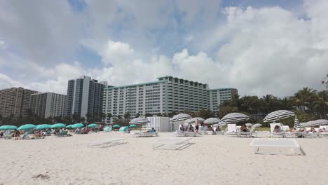 Menschen-Entspannen-Unter-Gestreiften-Sonnenschirmen-An-Einem-Sonnigen-Strand-In-Miami-Mit-Hotels-Im-Hintergrund