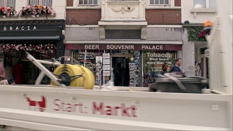 Storefront-Of-Souvenir-Shop-In-Bruges,-Belgium-With-Pedestrians-And-Vehicles-Passing-By-In-Foreground