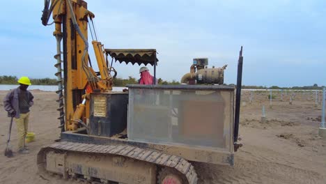 Panning-view-of-pile-driver-rig-machine-digging-for-solar-farm-concrete-pile-construction-on-dry-dusty-project-site