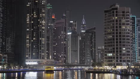 Glide-along-Dubai-Marina's-water-canal,-capturing-the-skyline-beneath-the-overhead-bridge-at-night