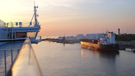 In-the-harbor-area,-a-ro-ro-ferry-is-slowly-moving-towards-a-red-black-cargo-ship-that-is-moored-in-a-beautiful-evening-sunset