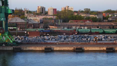 Piedras,-Piedras-Trituradas-Diversas,-Vagones-De-Tren,-Grúas-Para-Levantar-Carga-Y-Pontones-Con-Equipos-De-Construcción-En-La-Zona-Del-Muelle-Del-Puerto.