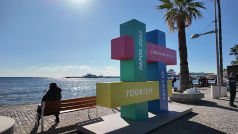 A-colorful-tourism-sign-for-Pafos-2023-stands-by-the-waterfront,-with-people-relaxing-on-benches-and-the-sea-in-the-background