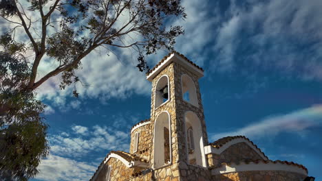 Bell-tower-and-stone-church-with-a-tree-under-a-partly-cloudy-sky