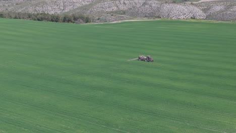 Vuelo-Con-Dron-En-Un-Campo-De-Cultivo-Extensivo-Donde-Nos-Dirigimos-A-Un-Tractor-Con-Remolque-Pulverizador-Donde-Está-Realizando-Trabajos-Agrícolas-En-El-Cultivo,-Se-Crean-Líneas-De-Las-Huellas-Del-Tractor