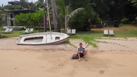 Hombre-Sentado-En-La-Playa-De-Ao-Tan-Al-Lado-De-Un-Barco-Rústico-Vacío