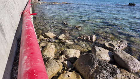 Close-up-of-a-rocky-shoreline-with-clear-water-in-Pafos,-Cyprus,-with-a-red-pipe-running-along-the-sea-wall