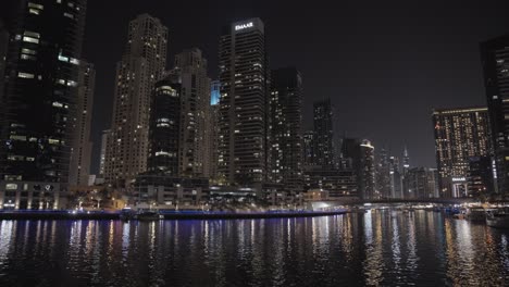 Crucero-Nocturno-Tranquilo-Por-El-Canal-Acuático-Del-Puerto-Deportivo-De-Dubai-Con-Un-Puente-Elevado-En-Foco