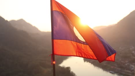 Laos-flag-flapping-in-the-wind-at-sunset-in-the-mountain-town-of-Nong-Khiaw-in-Laos,-Southeast-Asia