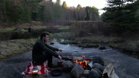 Hombre-Cocinando-Sobre-Una-Fogata-En-Un-Bosque-Cerca-De-Un-Arroyo,-Rodeado-De-Naturaleza-Con-Una-Linterna-Y-Una-Caña-De-Pescar-Cerca