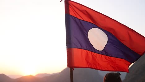 Bandera-De-Laos-Ondeando-Al-Viento-Al-Atardecer-En-La-Ciudad-Montañosa-De-Nong-Khiaw-En-Laos,-Sudeste-Asiático