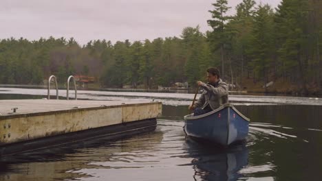 A-man-paddles-a-blue-canoe-near-a-wooden-dock-on-a-calm-lake,-surrounded-by-tall-pine-trees-and-a-tranquil-natural-landscape,-under-an-overcast-sky