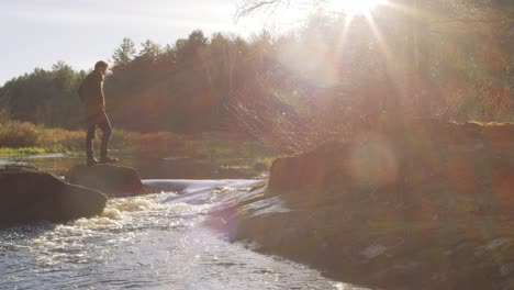 Man-standing-on-a-rock-by-a-river-in-a-forest-at-sunrise,-surrounded-by-nature-and-bathed-in-morning-light