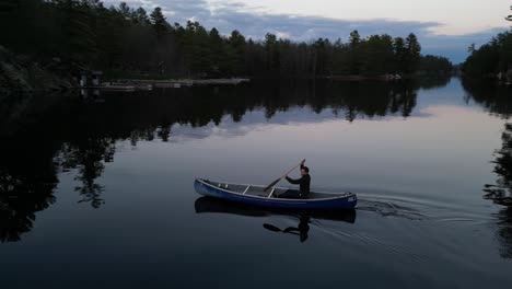 Persona-Navegando-En-Canoa-Por-Un-Lago-Sereno-Al-Atardecer,-Con-Reflejos-De-árboles-En-Las-Aguas-Tranquilas-Y-Una-Atmósfera-Pacífica