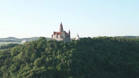 Majestic-historical-castle-atop-a-verdant-hill,-encircled-by-a-dense-forest-under-a-clear-sky