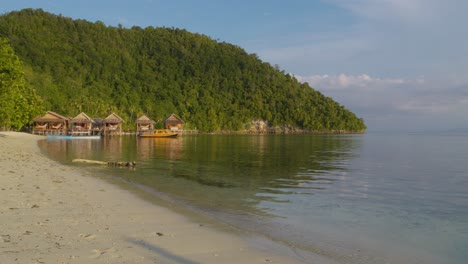 Tranquil-beach-on-Kri-Island-in-the-Raja-Ampat-Archipelago,-Indonesia,-featuring-wooden-huts-along-the-shore-against-a-lush-green-backdrop