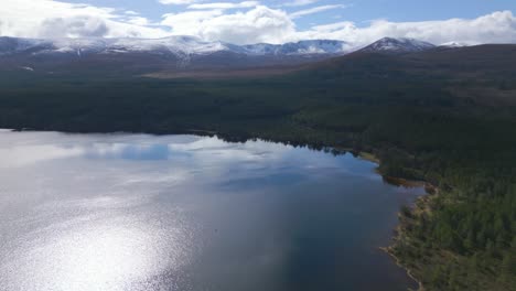 Aerial-rising-from-Loch-Morlich,-snow-capped-mountains-in-background,-Scotland