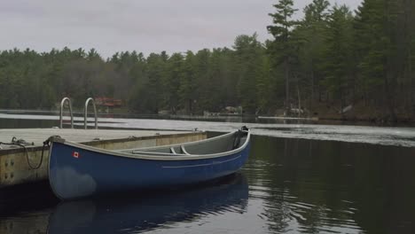 Una-Canoa-Azul-Está-Atracada-En-Un-Muelle-De-Madera-En-Un-Lago-Tranquilo,-Rodeada-De-Altos-Pinos-Y-Un-Sereno-Paisaje-Natural,-Bajo-Un-Cielo-Nublado