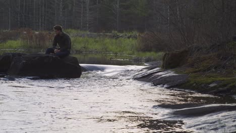 Man-sitting-on-a-rock-by-a-forest-river,-lost-in-contemplation,-surrounded-by-nature-and-flowing-water