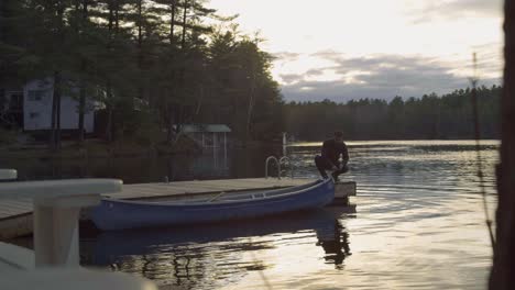 Ein-Mann-Steht-Bei-Sonnenuntergang-Auf-Einem-Holzsteg-Neben-Einem-Blauen-Kanu