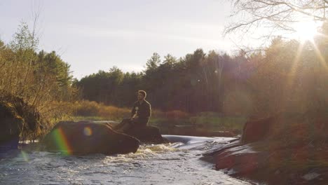 Man-sitting-on-a-rock-by-a-river-in-a-forest-at-sunset,-surrounded-by-trees-and-bathed-in-golden-light